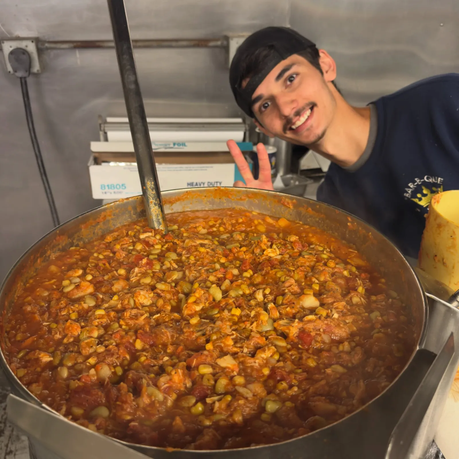 A BBQ King cook throwing a peace sign beside a full kettle of fresh Brunswick stew