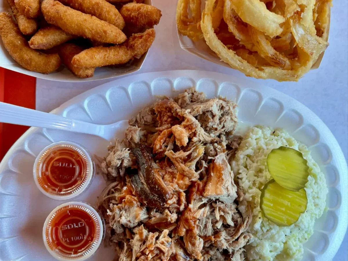 Overhead BBQ plate on a red tray with chopped pit-smoked pork, white slaw and pickles, hushpuppies, and onion rings